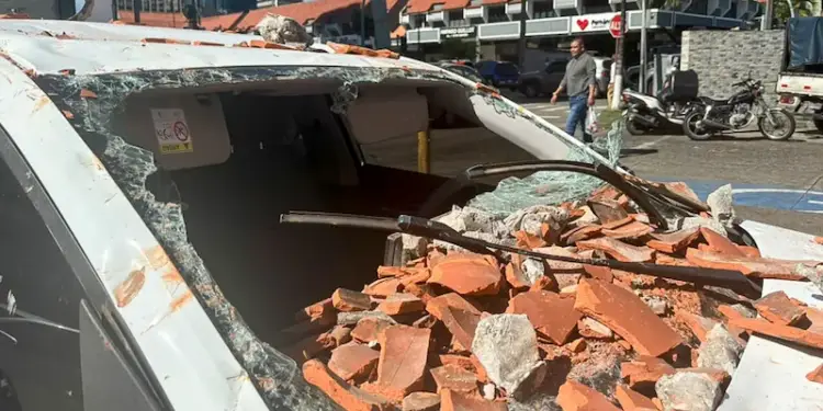Coche blanco gravemente dañado por escombros de ladrillos y hormigón sobre el parabrisas y techo, en una calle urbana.