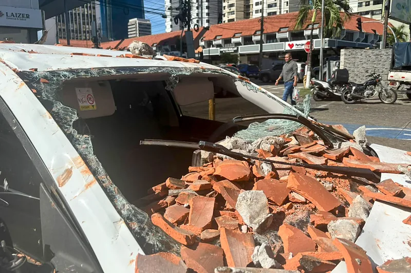 Coche blanco gravemente dañado por escombros de ladrillos y hormigón sobre el parabrisas y techo, en una calle urbana.