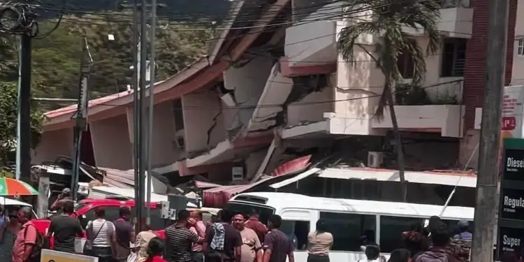 Edificio de varios pisos con colapso parcial y daños estructurales. Personas observan la escena. Señal de 'PELIGRO'. Cables y montañas.