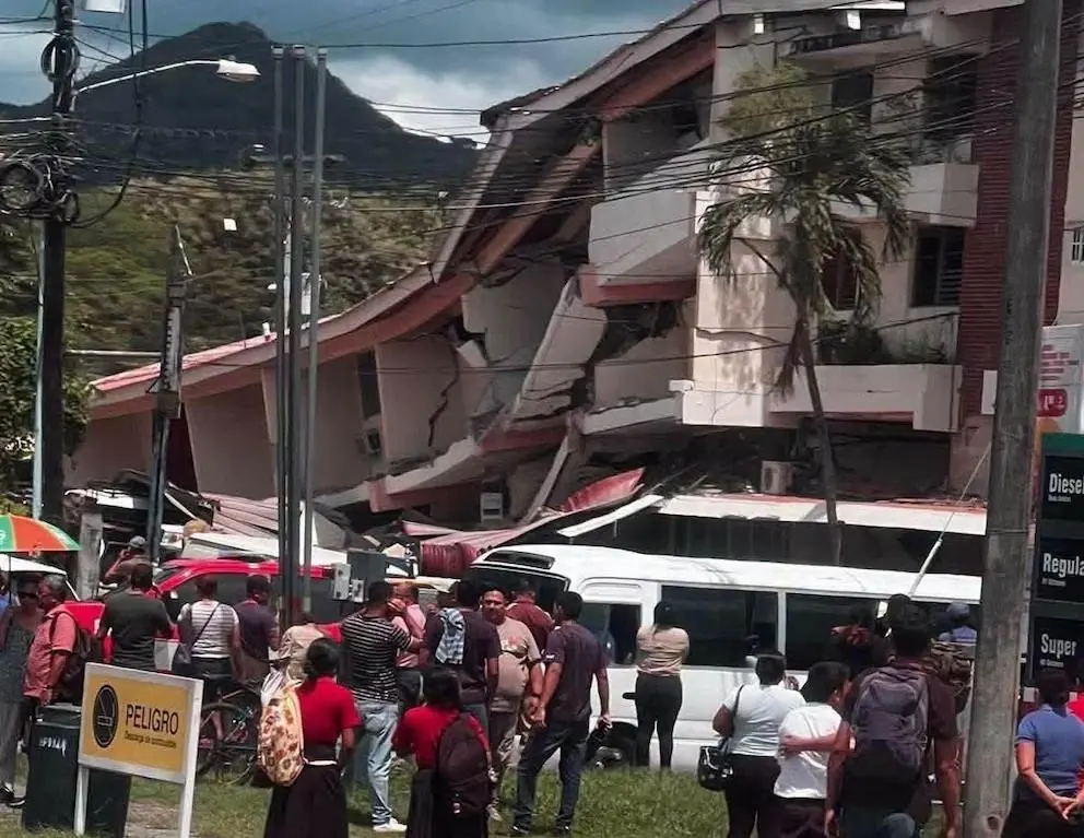 Edificio de varios pisos con colapso parcial y daños estructurales. Personas observan la escena. Señal de 'PELIGRO'. Cables y montañas.