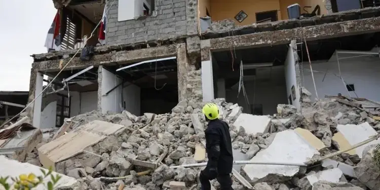 Un rescatista con casco amarillo y traje oscuro camina entre los escombros de un edificio de varios pisos parcialmente destruido por un desastre.