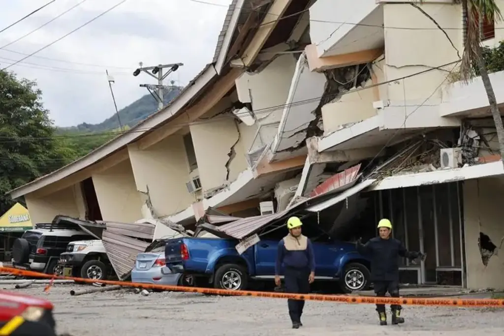 Edificio colapsado y vehículos dañados tras un desastre natural, con personal de rescate enfrente.