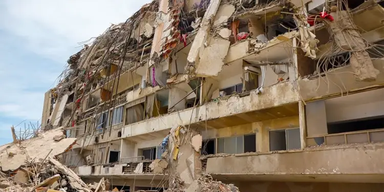 Edificio de varios pisos gravemente derrumbado con escombros, hormigón roto y barras de refuerzo expuestas. Gran pila de ruinas en la base.