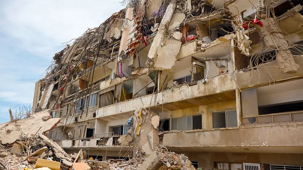 Edificio de varios pisos gravemente derrumbado con escombros, hormigón roto y barras de refuerzo expuestas. Gran pila de ruinas en la base.