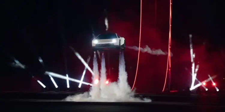 Un vehículo blanco de diseño angular y futurista flota en el aire, impulsado por chorros de humo blanco, bajo luces de escenario rojas y blancas.