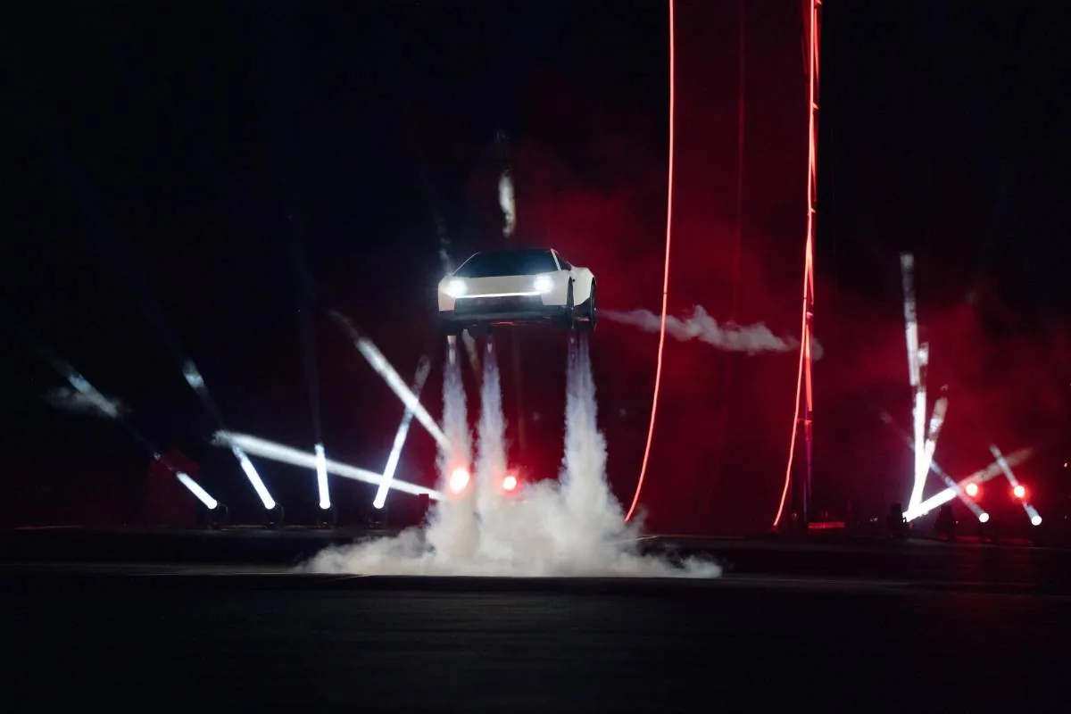Un vehículo blanco de diseño angular y futurista flota en el aire, impulsado por chorros de humo blanco, bajo luces de escenario rojas y blancas.