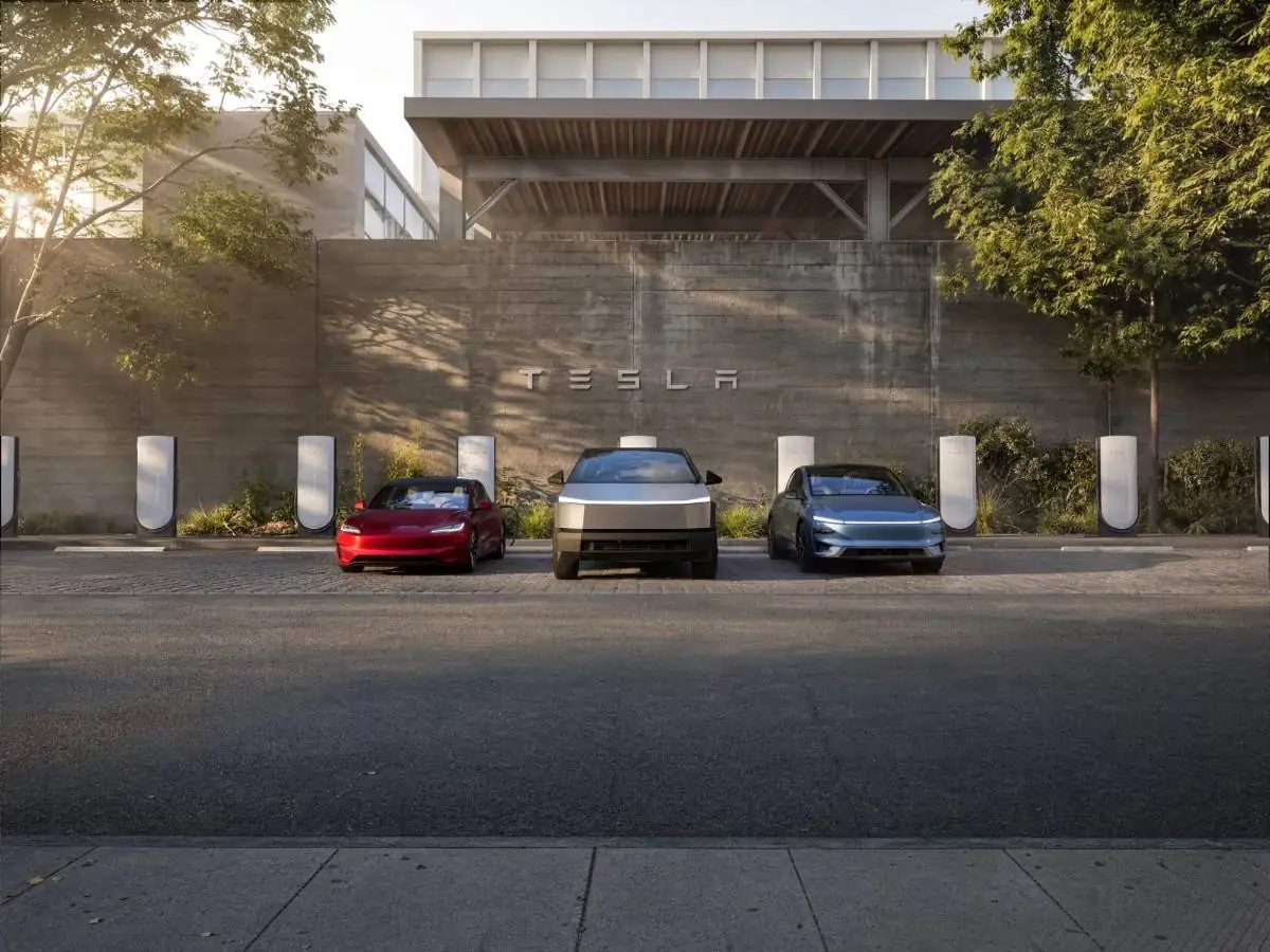 Tres vehículos eléctricos estacionados frente a una pared de concreto con el logotipo de Tesla y múltiples estaciones de carga.