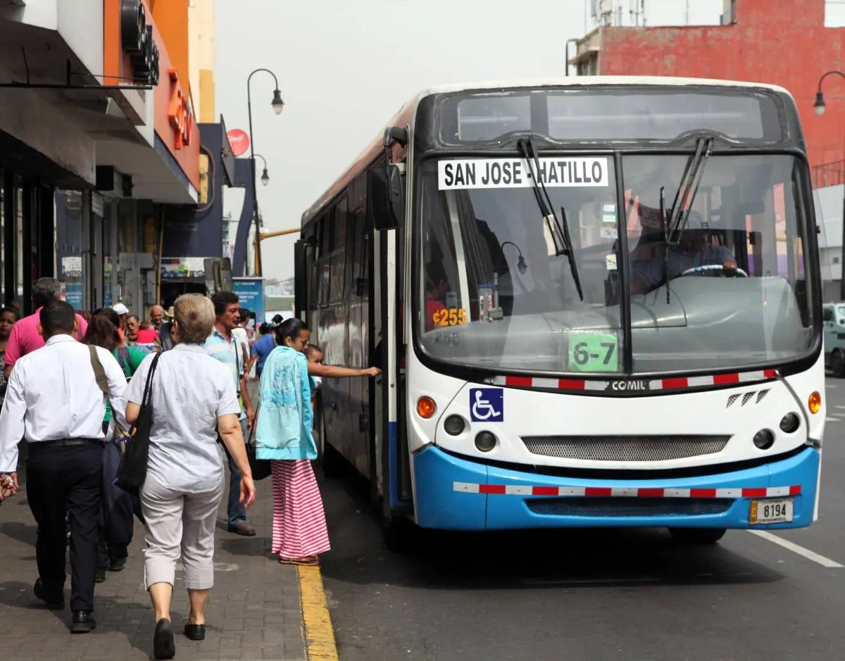 Autobús urbano detenido en la calle mientras personas caminan por la acera y una mujer aborda el vehículo.