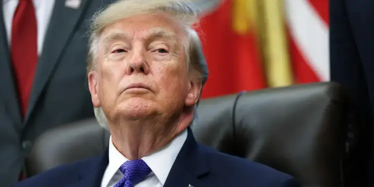 Retrato de Donald Trump sentado con traje azul y corbata morada frente a banderas
