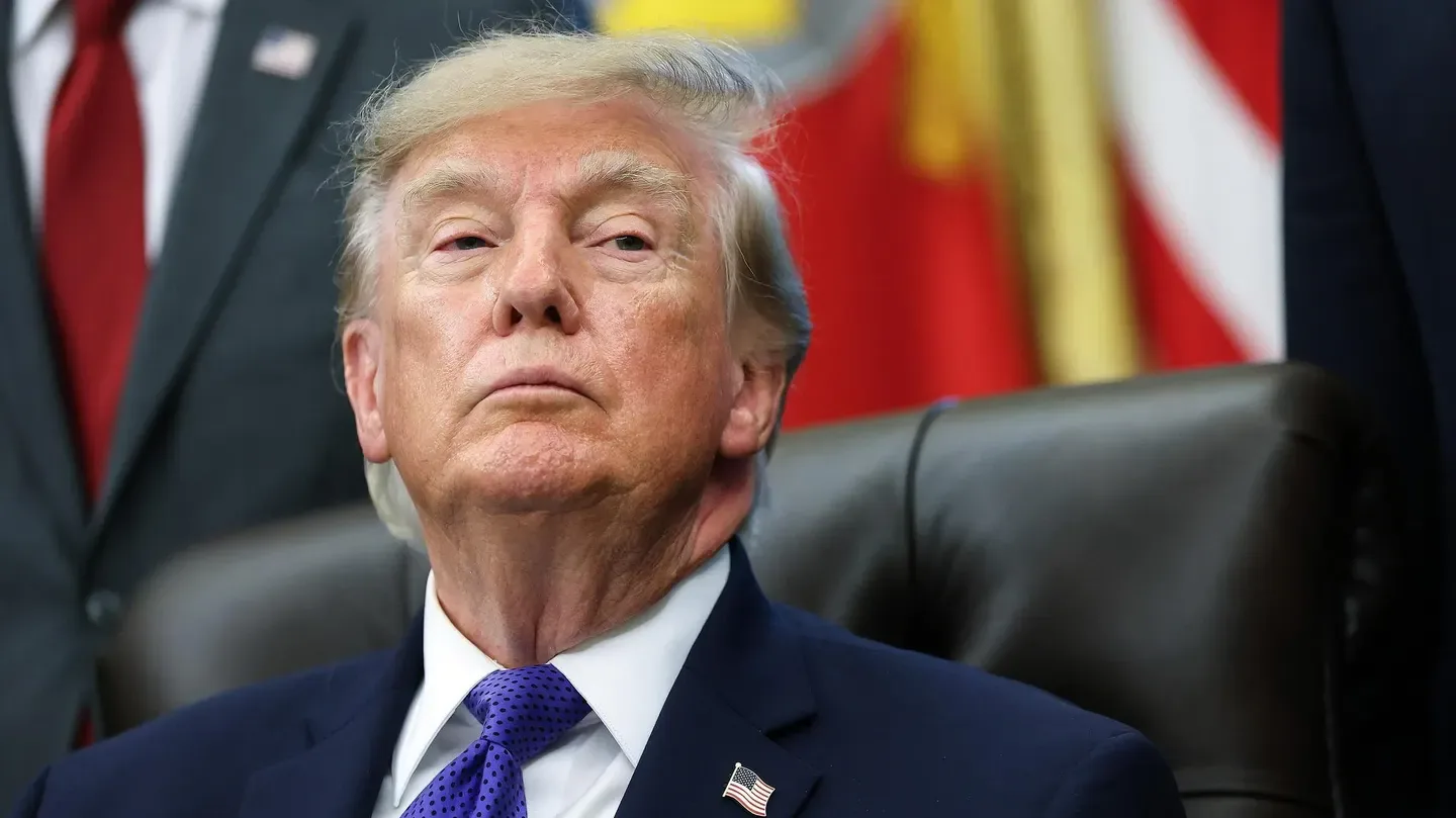 Retrato de Donald Trump sentado con traje azul y corbata morada frente a banderas