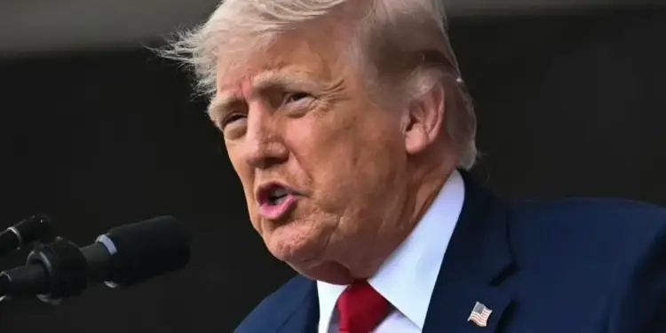 Retrato de medio cuerpo de Donald Trump hablando ante un micrófono con traje azul y corbata roja.
