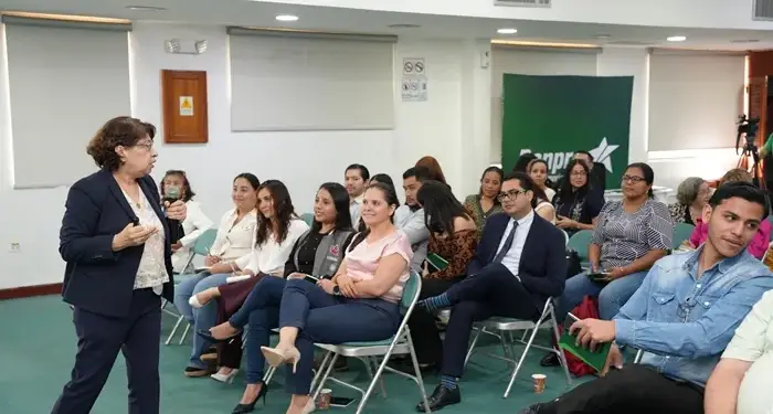 Mujer hablando con micrófono frente a una audiencia atenta en una sala de conferencias corporativa.
