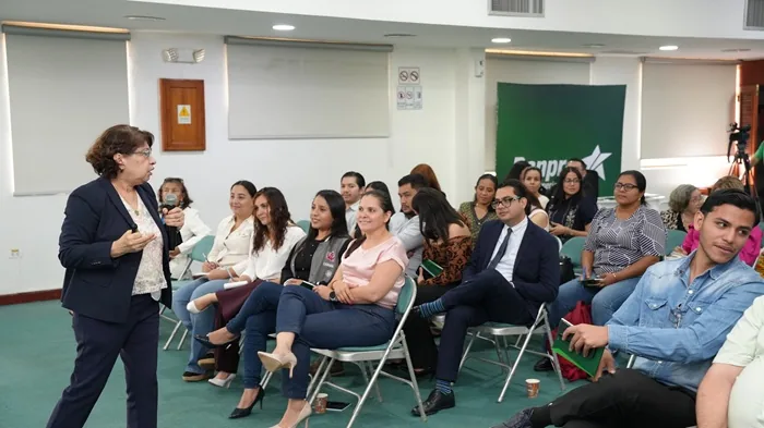 Mujer hablando con micrófono frente a una audiencia atenta en una sala de conferencias corporativa.