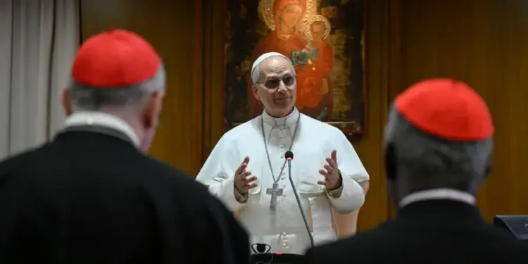 Un líder religioso con vestiduras blancas hablando frente a dos cardenales con solideos rojos.