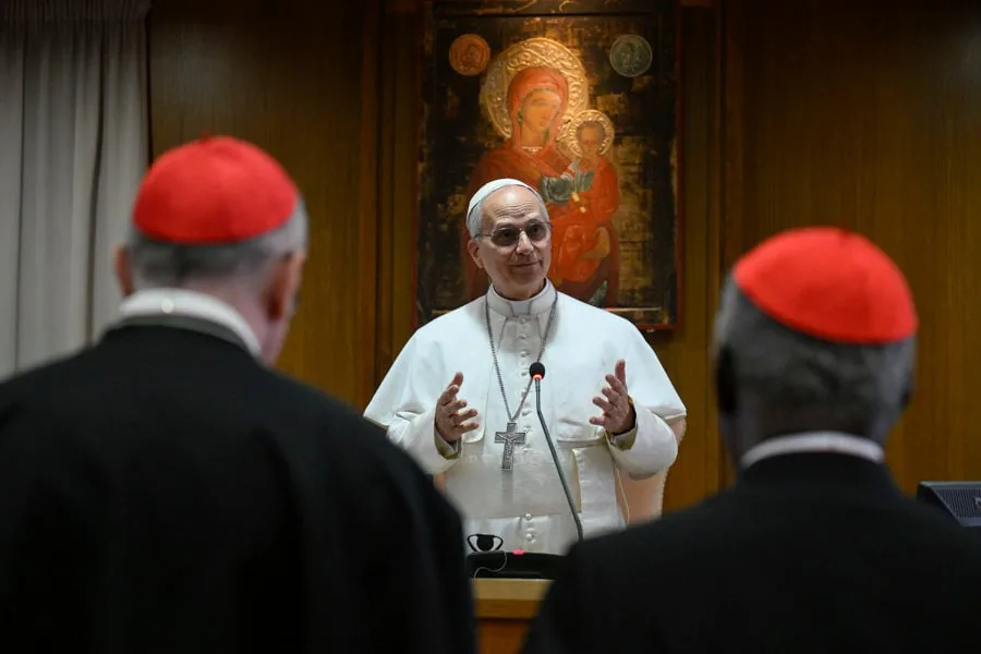 Un líder religioso con vestiduras blancas hablando frente a dos cardenales con solideos rojos.