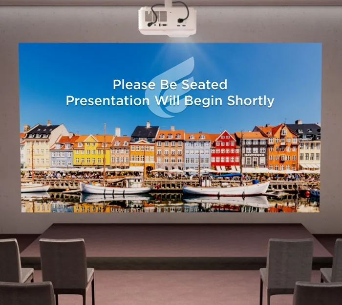 Proyector en sala de conferencias mostrando mensaje 'Please Be Seated Presentation Will Begin Shortly' sobre imagen de puerto con edificios coloridos y barcos. Asientos vacíos.