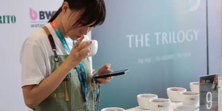 Mujer con delantal verde probando café y usando su teléfono en un evento profesional.